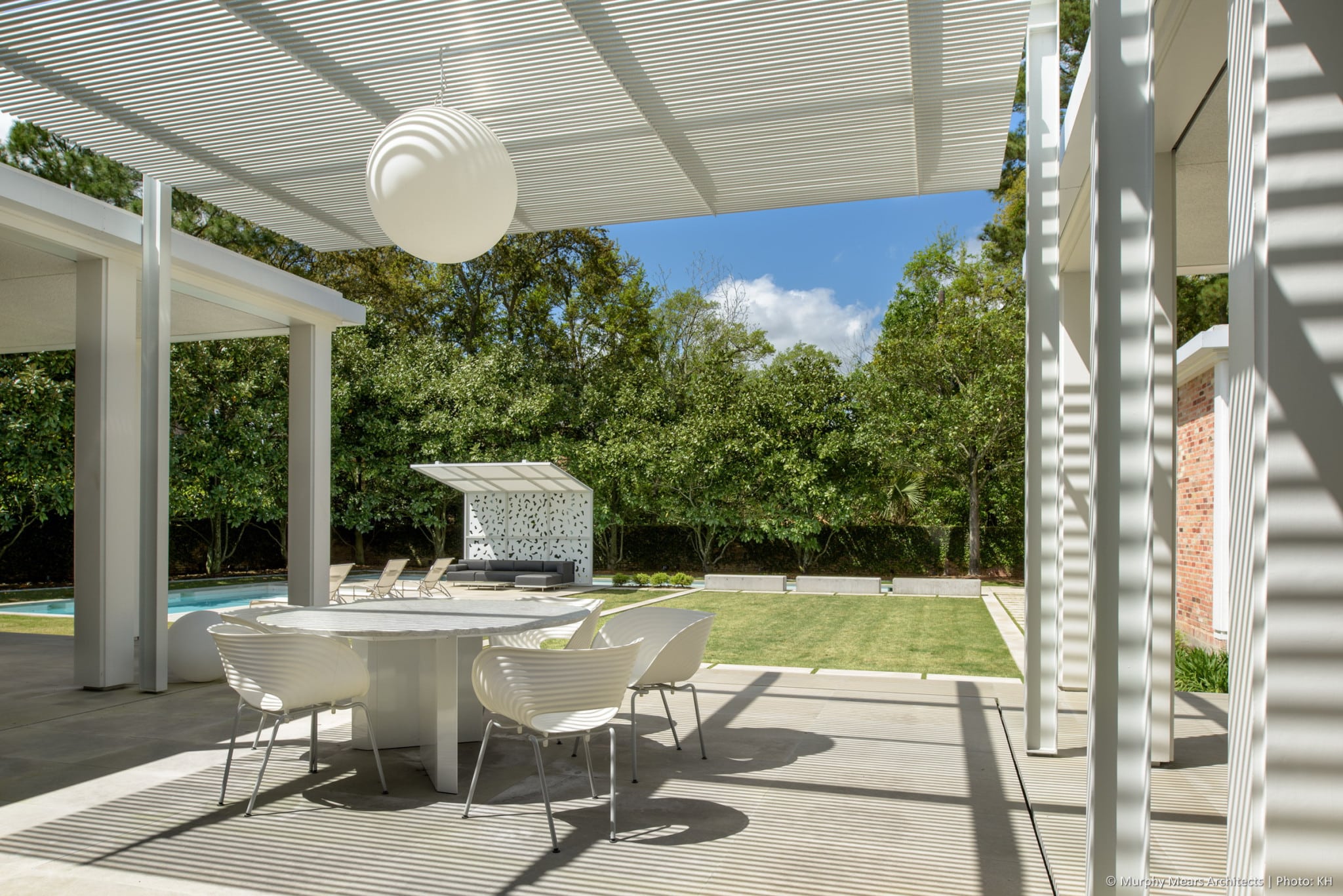 Outdoor dining area beneath the aluminum and steel trellis, overlooking the central lawn, with the pool terrace shade canopy beyond.